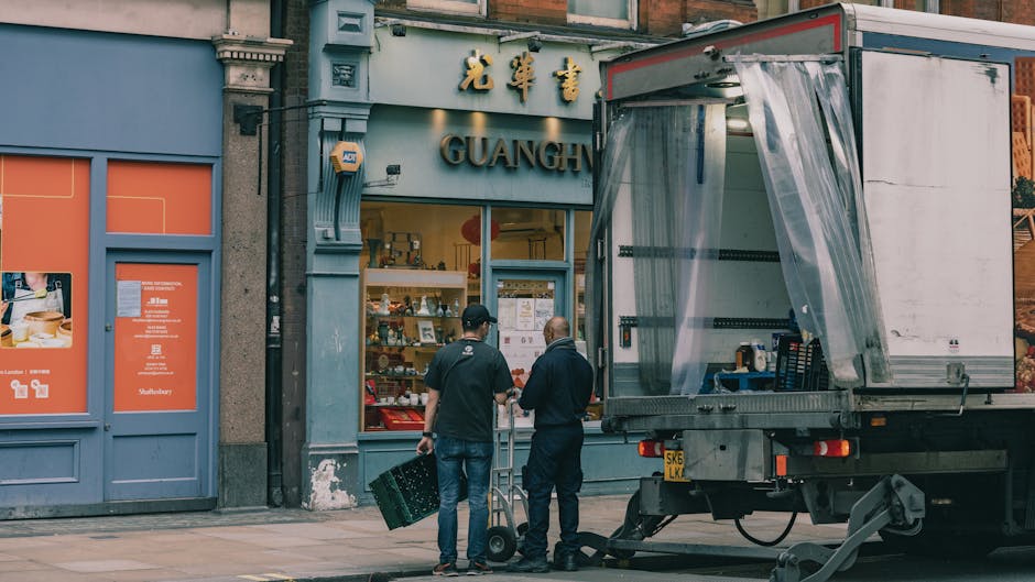 Two removal workers from South Kensington Man and Van are preparing for a home relocation outside a shop with a blue façade and gold signage that reads 'GUANG' in an urban area. The workers, dressed i