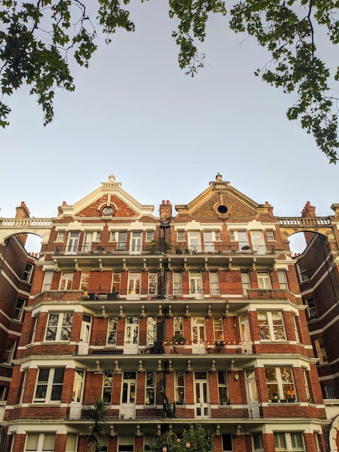 A rectangular white metal sign with black and red lettering that reads 'Welcome to South Kensington SW7' is mounted on a red-brown brick wall. The sign is positioned horizontally and surrounded by vis