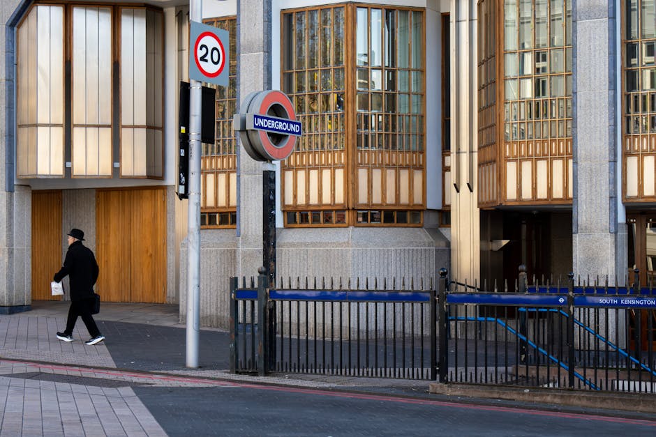 A man dressed in dark clothing and a black hat is walking along a city sidewalk outside a modern building with large glass windows framed by wooden panels. In front of the building, there is a black metal fence with a sign indicating 'South Kensington' and a blue railing, suggesting a restricted pedestrian area or entrance. A street sign indicates a 20 mph speed limit, and an 'Underground' roundel sign marks the nearby South Kensington tube station. The scene is well-lit with natural daylight, and the sidewalk appears clean and wide, suitable for residential or commercial activities. The area visible is part of an urban environment involved in regular pedestrian movement, possibly close to property entrances or vehicle access points used during home relocation or furniture transport operations; these elements are typical in areas served by removals and moving services like those offered by South Kensington Man and Van.