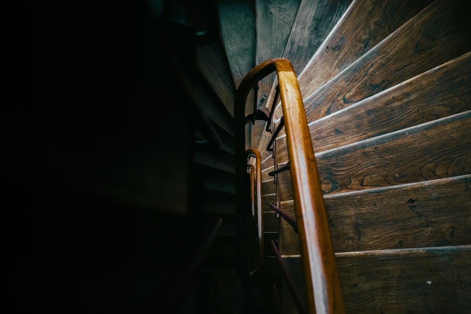 A narrow outdoor staircase leading down from a house, with stone steps and a black metal railing on one side. The area is enclosed by an exterior wall with a small window framed in red and a white dow
