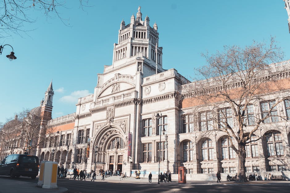 A view of the exterior of the Victoria and Albert Museum in South Kensington with a clear sky overhead. In the foreground, a paved pathway and a grassy area are visible, leading towards the museum ent